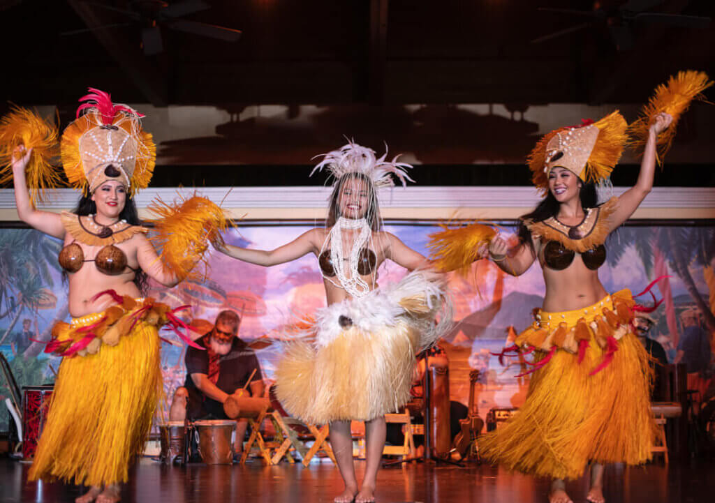 Three hula dancers smiling as they dance on a stage with musicians behind them.